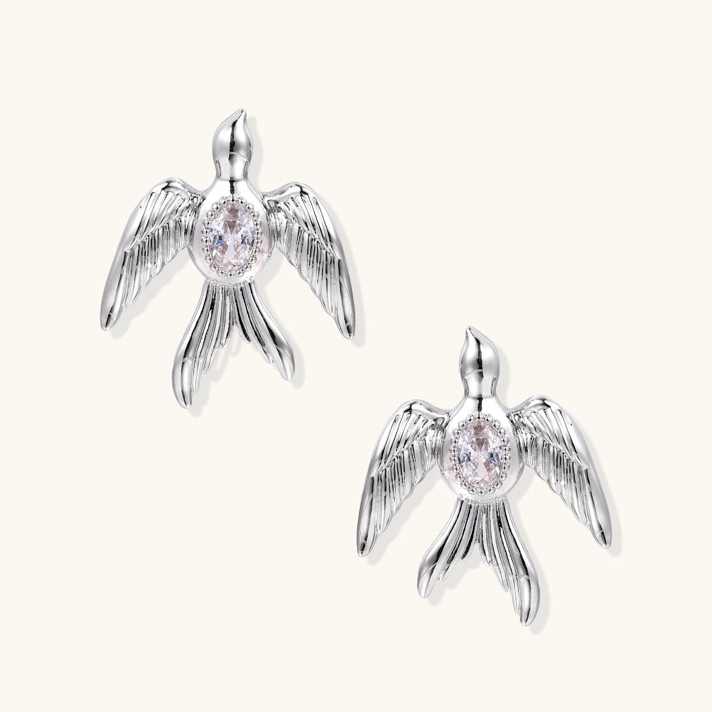 White gold-plated bird earrings with spread wings and oval zirconia center on cream background