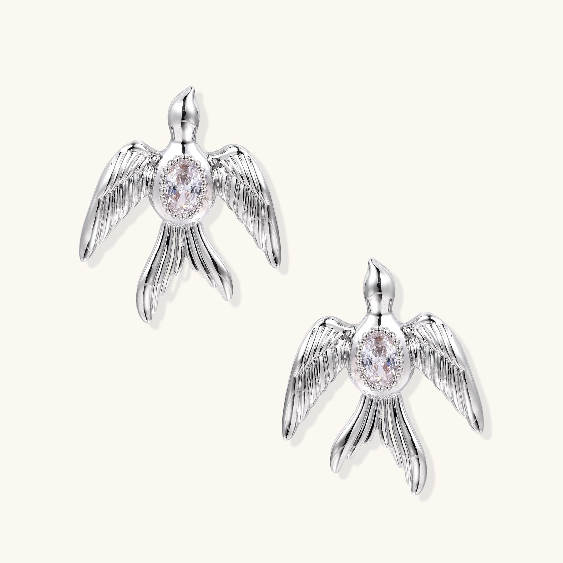 White gold-plated bird earrings with spread wings and oval zirconia center on cream background