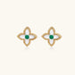 Gold quatrefoil stud earrings with white mother-of-pearl petals and emerald green gemstone center on beige background.