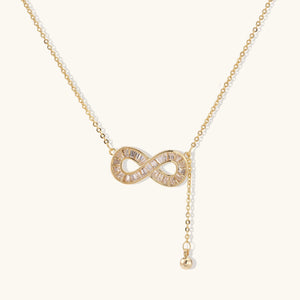 Gold infinity pendant with baguette crystals on adjustable chain against cream background.
