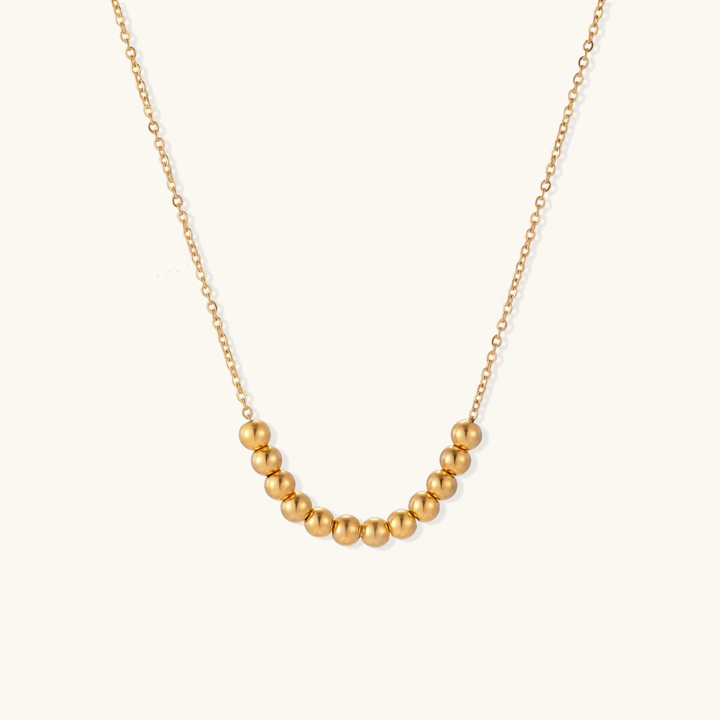 Delicate gold-plated necklace with graduated spherical beads forming gentle smile curve on beige background
