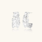 White gold plated sculptural hoop earrings with organic wavy edges and reflective metallic finish on cream background