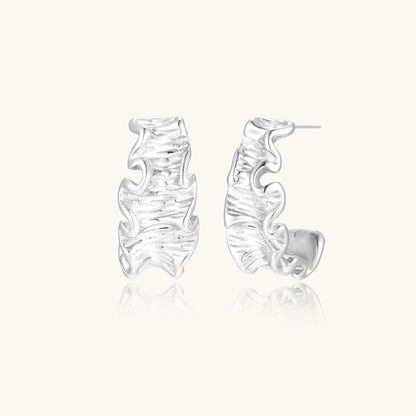 White gold plated sculptural hoop earrings with organic wavy edges and reflective metallic finish on cream background