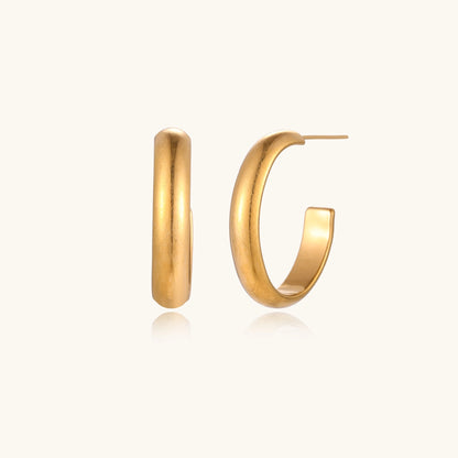 Chunky tubular hoop earrings in warm 14K gold-plated finish with smooth polished surface on cream background.