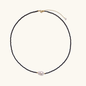 Delicate necklace with black blue sandstone beads and baroque pearl center on white background with gold accents.